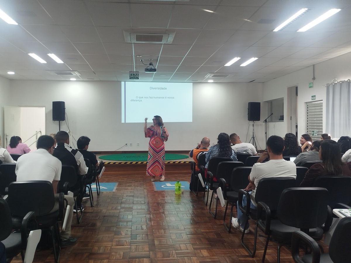 A psic&oacute;loga Vera L&uacute;cia de Matos Rodrigues, vestindo um longo estampado colorido, est&aacute; em p&eacute; no palco e fala para a plateia de servidores sentados em cadeiras no audit&oacute;rio.